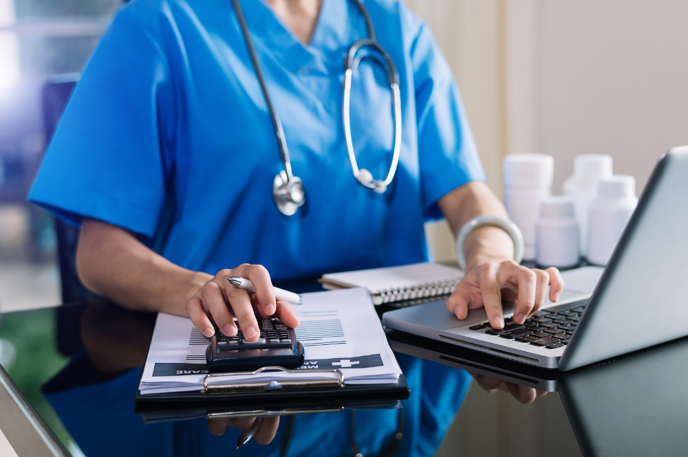 A woman in scrubs sits at a desk with a laptop and calculator, analyzing healthcare costs in retirement planning.