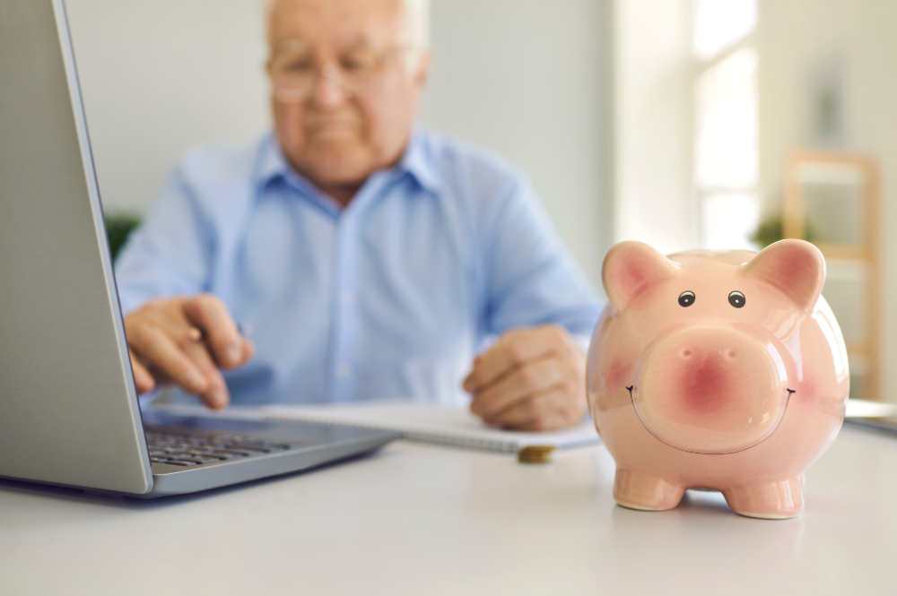 A person in a blue shirt uses a laptop at a desk, with a smiling piggy bank prominently displayed nearby, symbolizing annuities for retirement income.