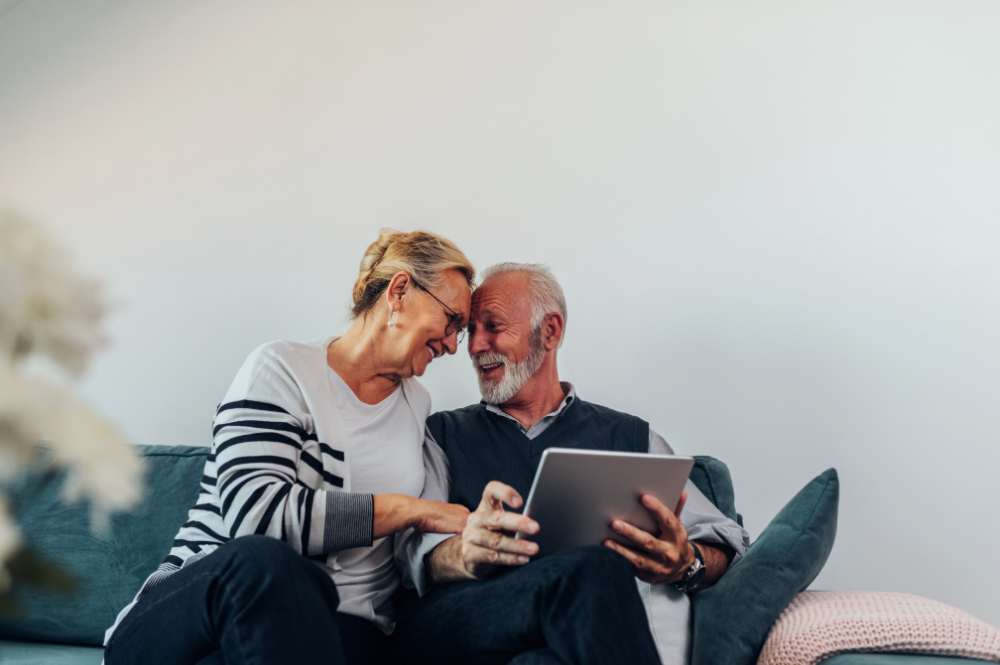 An older couple in their 50s on a couch, reviewing financial planning for retirement on a tablet.